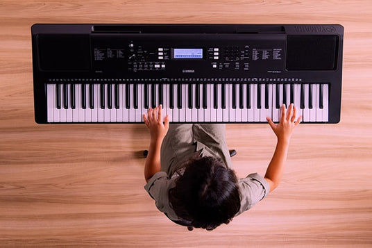 Top-down view of a person playing a black digital piano, with visible sheet music stand, electronic control panel, and full keyboard.