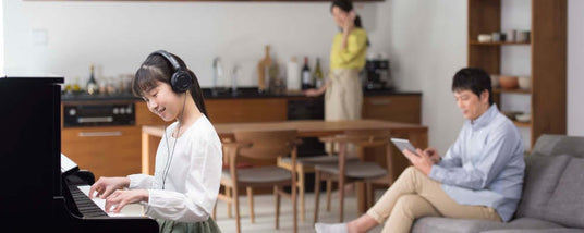 Young woman practicing piano with headphones in a home setting while a man seated on a sofa uses a tablet and another person stands in the background, indicating a quiet environment for focused music practice.
